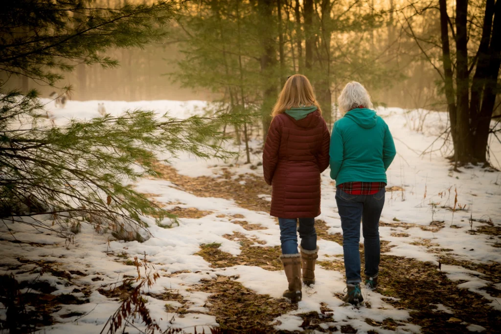 Two women, one older with gray hair and one younger with long hair, walk side by side on a snowy, wooded trail at sunset. As they stroll, they discuss estate planning misconceptions, bundled in winter jackets and boots amid trees and patches of snow.