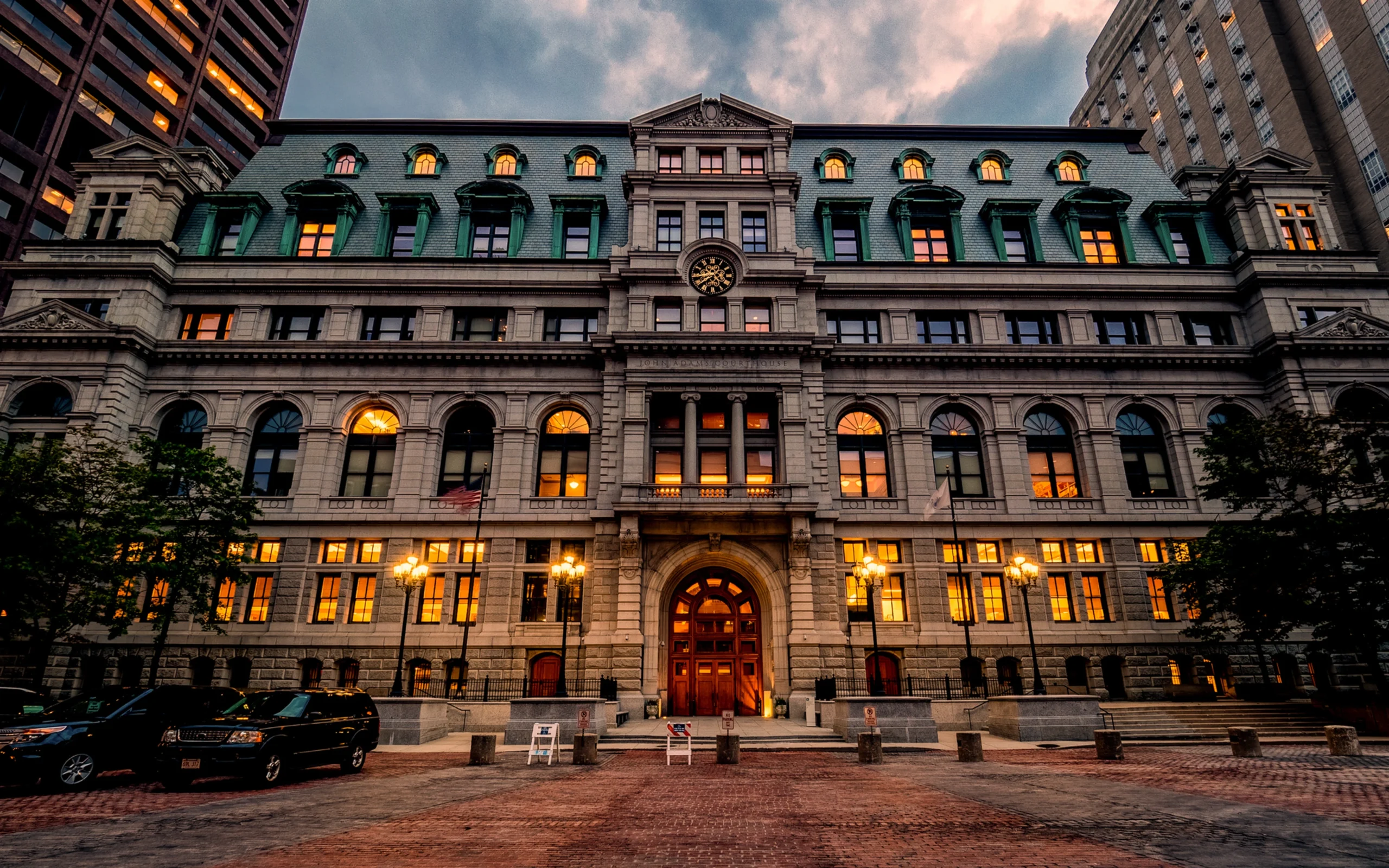 A grand, historic building with tall arched windows and a central clock is lit warmly at dusk. Cars are parked in front, evoking the importance of choosing guardians, executors, and trustees under a dramatic, clouded sky.