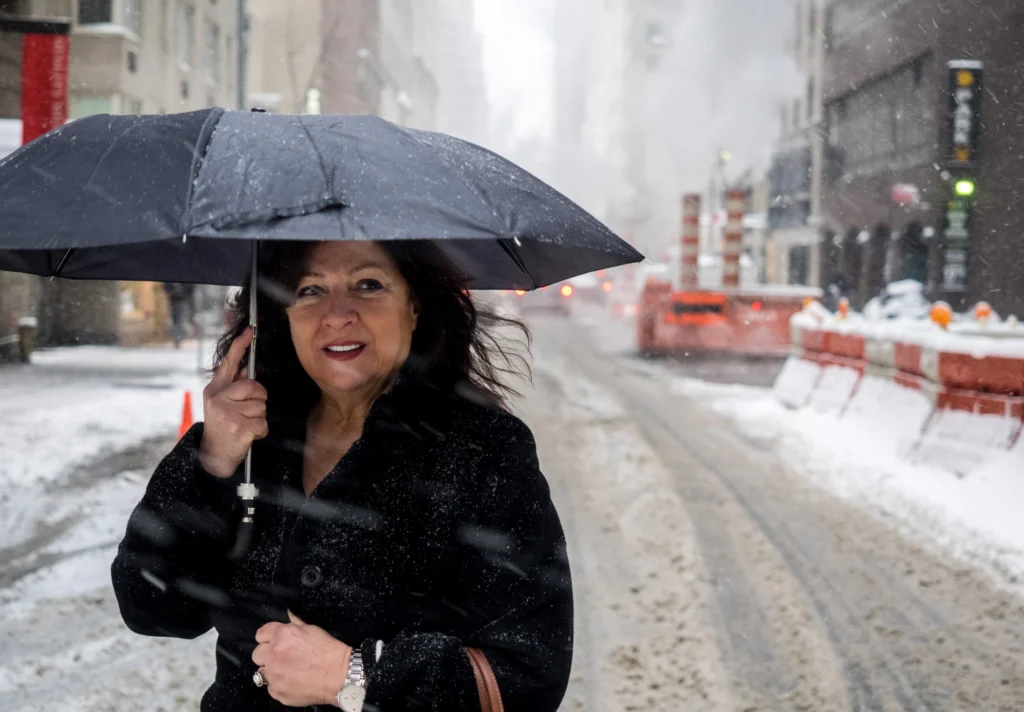 A woman in a black coat holds a black umbrella, standing on a snowy city street lined with orange barricades and tall buildings. She is smiling at the camera, perhaps considering the benefits of revocable trusts for her future planning.