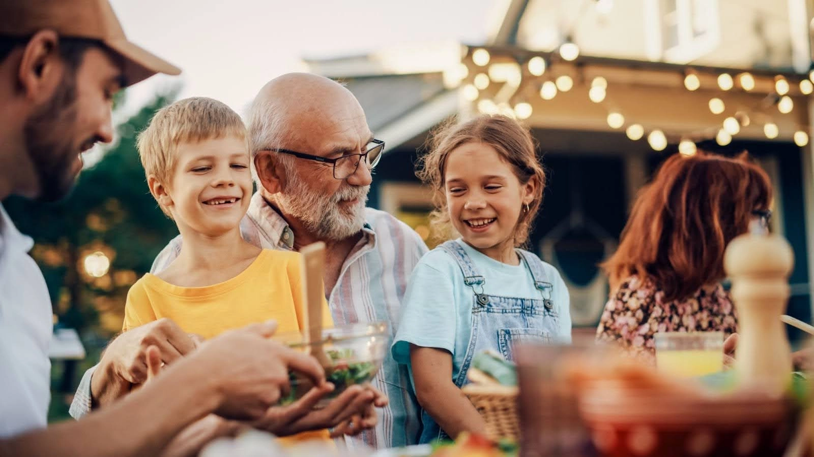 A smiling elderly man sits outdoors with a young boy and girl, sharing a meal. Nearby people and glowing string lights create a warm, festive atmosphere—perfect for discussing family matters like probate vs non-probate assets in Massachusetts.