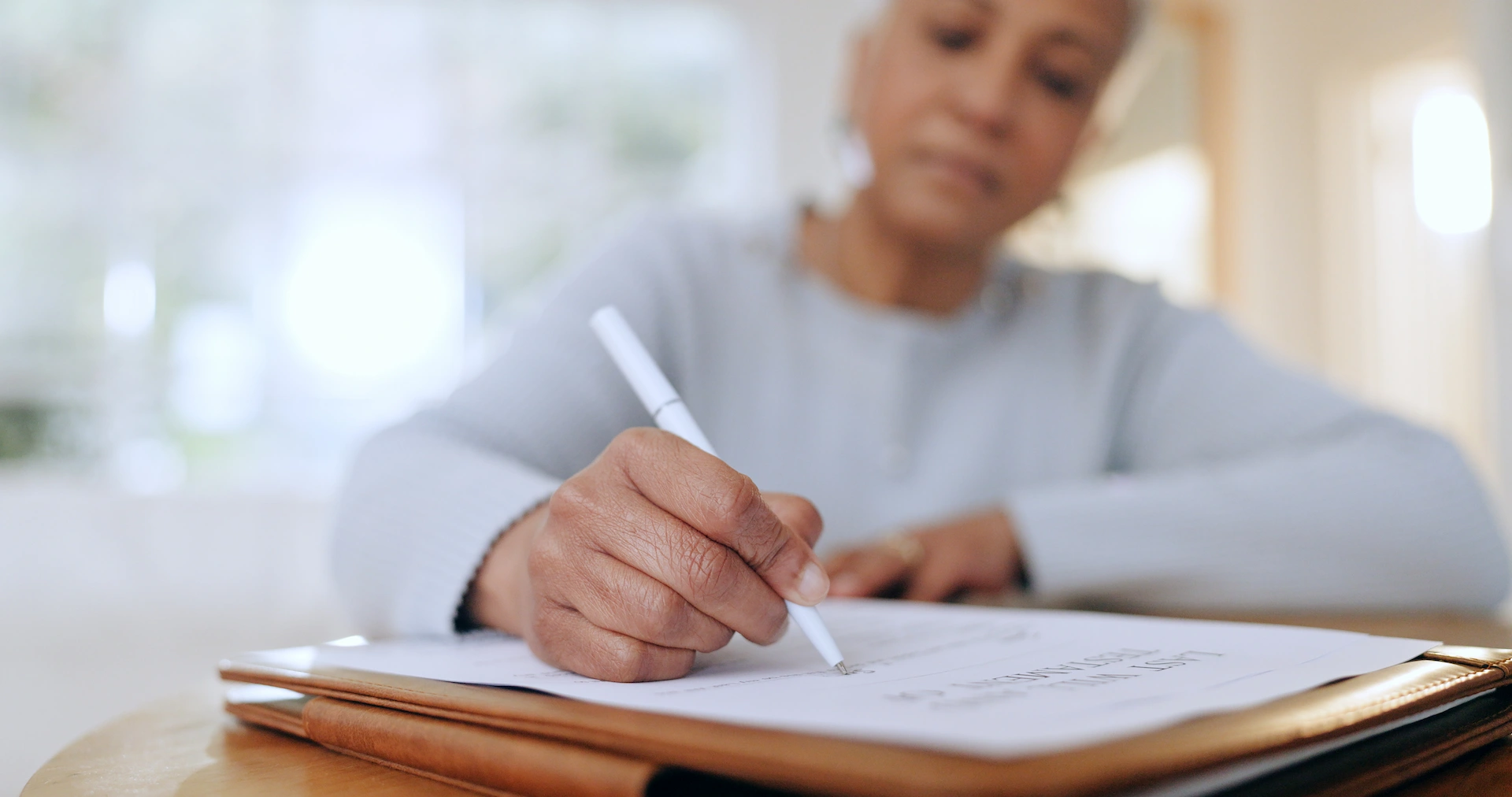 A person sits at a table, holding a pen and writing on a document placed on a clipboard. The focus is on their hand and the paper, with their face softly blurred in the background.