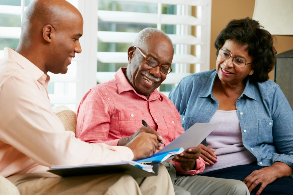 Three adults sit together indoors. A man in a pink shirt smiles as he signs a document, assisted by a younger man holding papers, while a woman in glasses and a blue shirt sits beside them, watching and smiling.