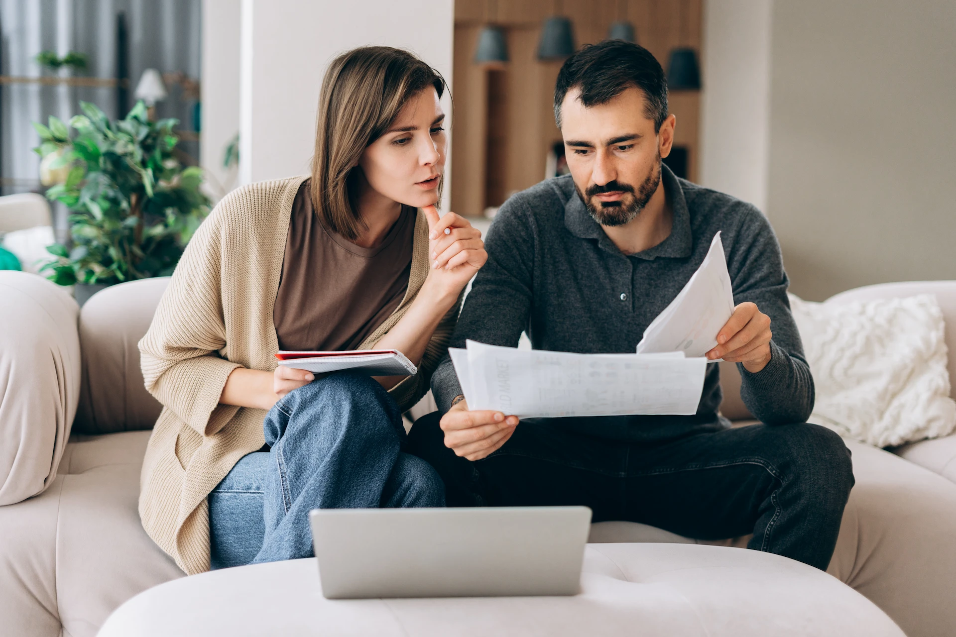 A woman and man sit on a couch looking thoughtfully at financial documents and a laptop, with the woman holding a notebook and pen, suggesting they are reviewing their finances or budgeting together at home.