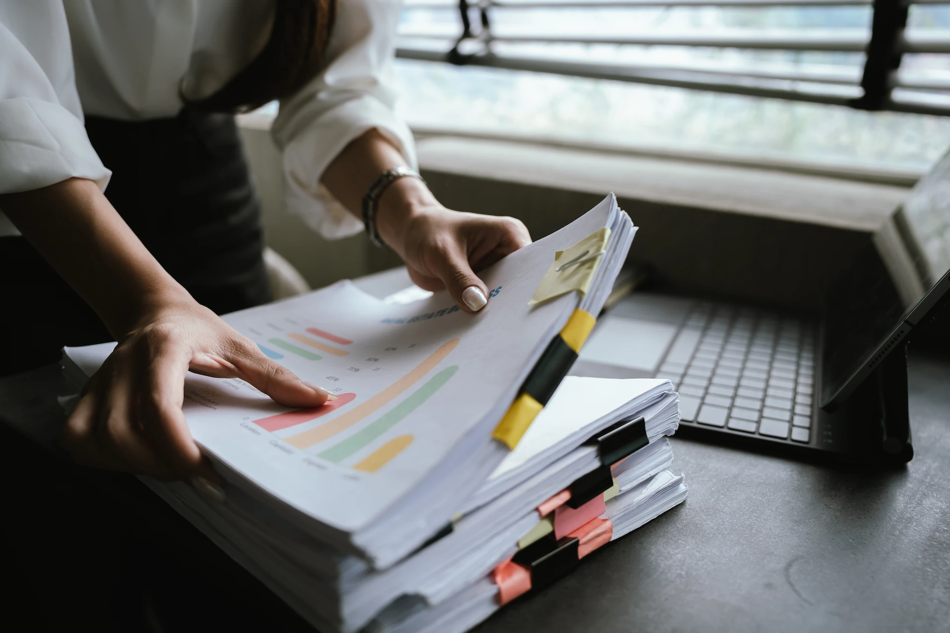 A person organizes a stack of printed documents with colorful tab markers on a desk near a laptop, with sunlight coming through window blinds in the background.