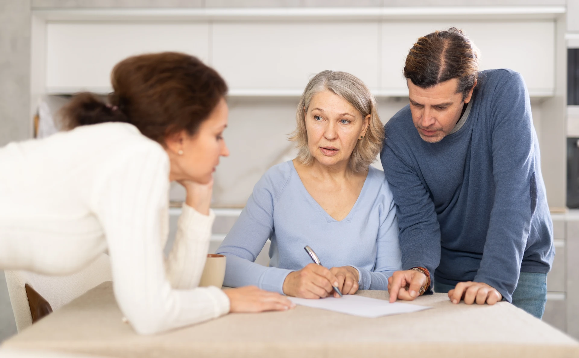 Three adults sit at a table, focused on paperwork. An older woman holds a pen, while a man beside her points at the paper and a younger woman leans in, listening attentively.