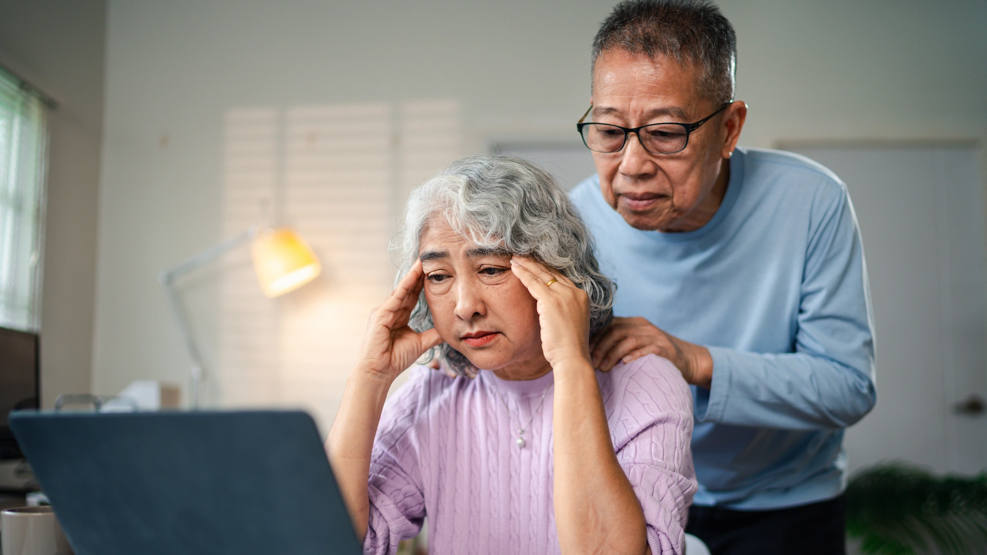 An older woman sits at a desk looking stressed, holding her temples, while an older man stands behind her with his hands on her shoulders. They both look at a laptop screen in a home setting.