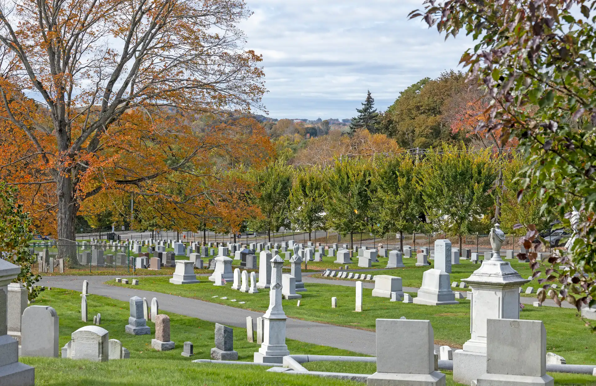 A peaceful cemetery with rows of gravestones and monuments set on green grass. Trees with autumn foliage surround the area under a cloudy sky, and a distant landscape is visible in the background.