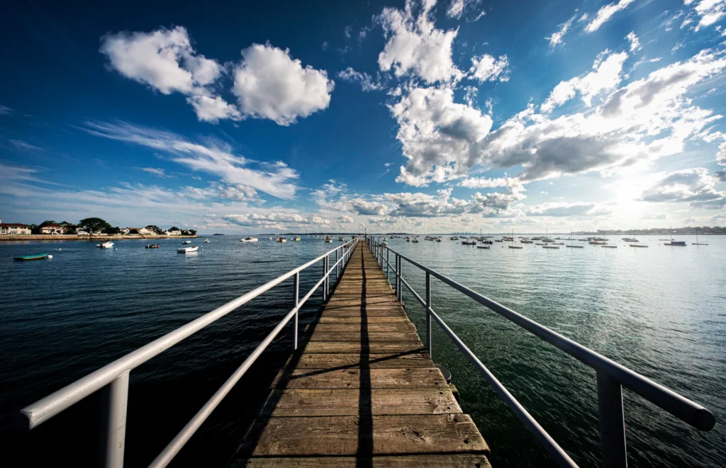 A wooden pier with railings extends over calm water toward the horizon, where small boats are anchored. The sky is bright blue with scattered clouds, and houses line the distant shoreline.