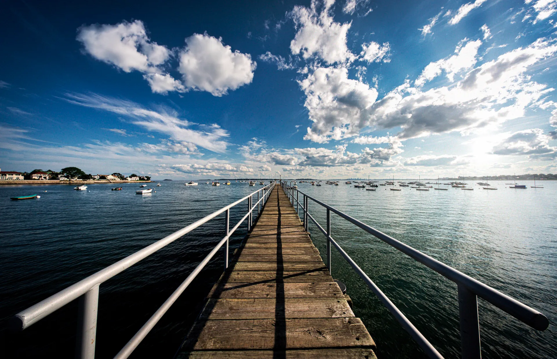 A wooden pier with railings extends over calm water toward the horizon, where small boats are anchored. The sky is bright blue with scattered clouds, and houses line the distant shoreline.