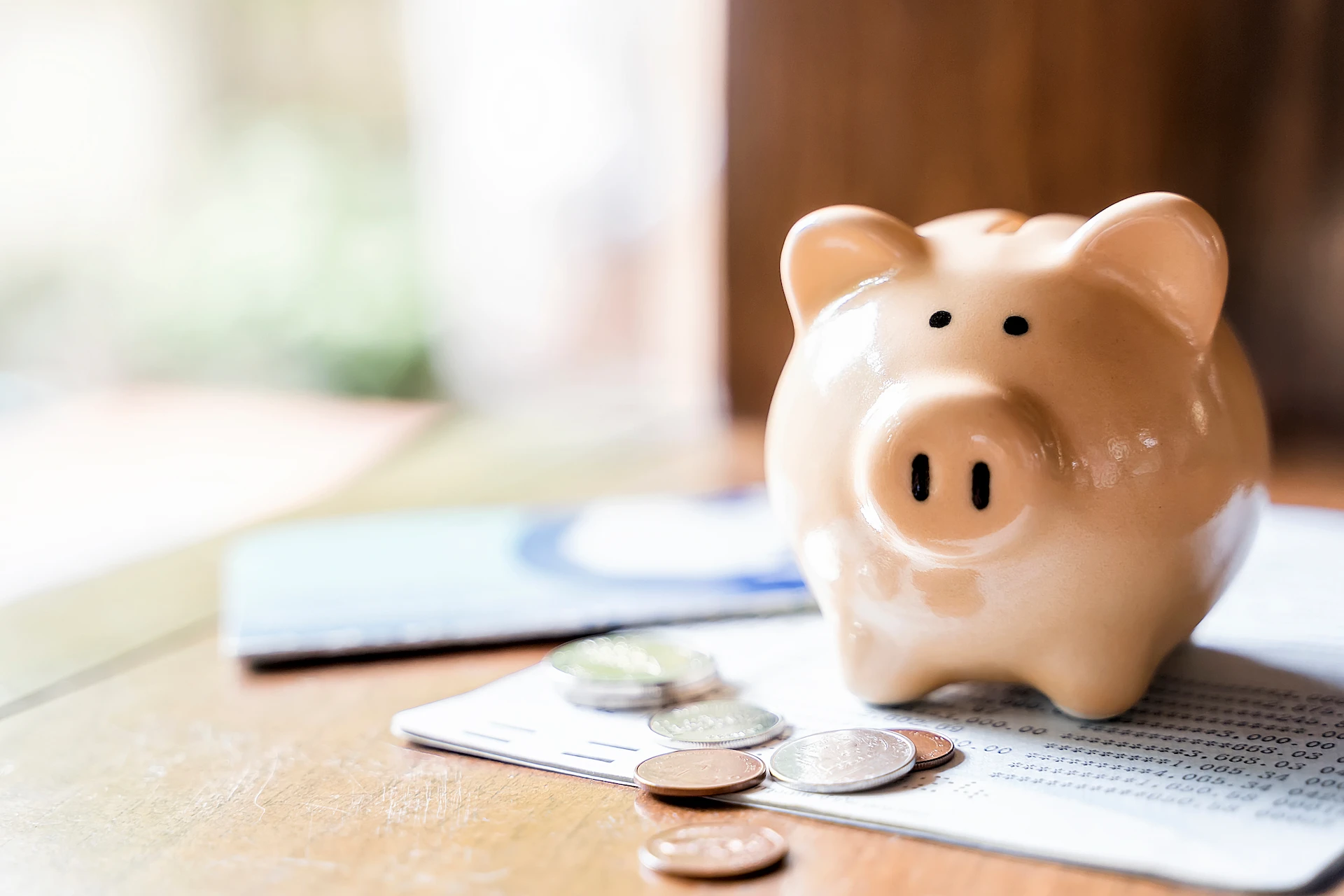 A beige piggy bank sits on a table next to coins, a bankbook, and a check card, symbolizing saving money and personal finance. The background is softly blurred.