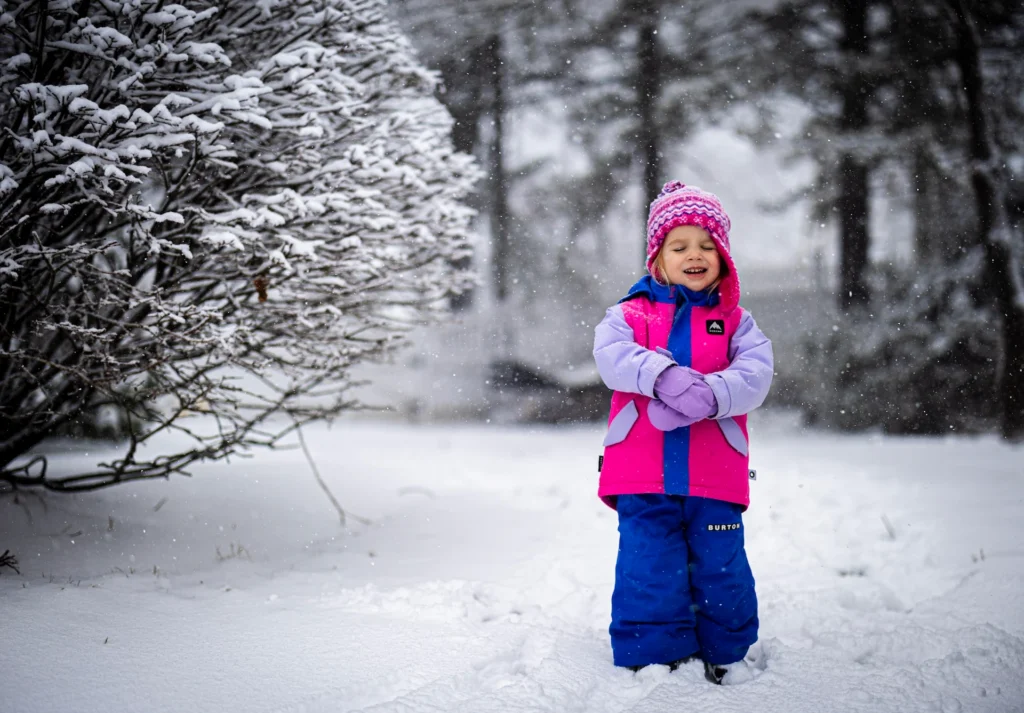 A young child wearing a pink hat, pink and purple winter coat, and blue snow pants stands smiling in a snowy outdoor scene, with snow-covered bushes and trees in the background. Snow is gently falling.