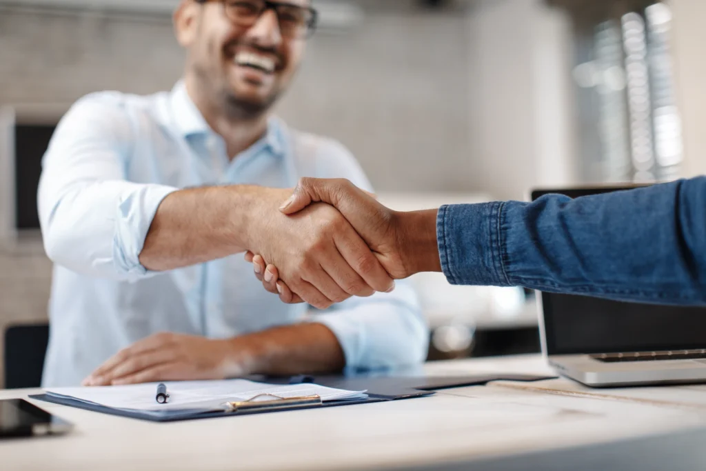 Two people shaking hands across a desk in an office setting, with one person smiling in the background. A clipboard and laptop are visible on the desk.