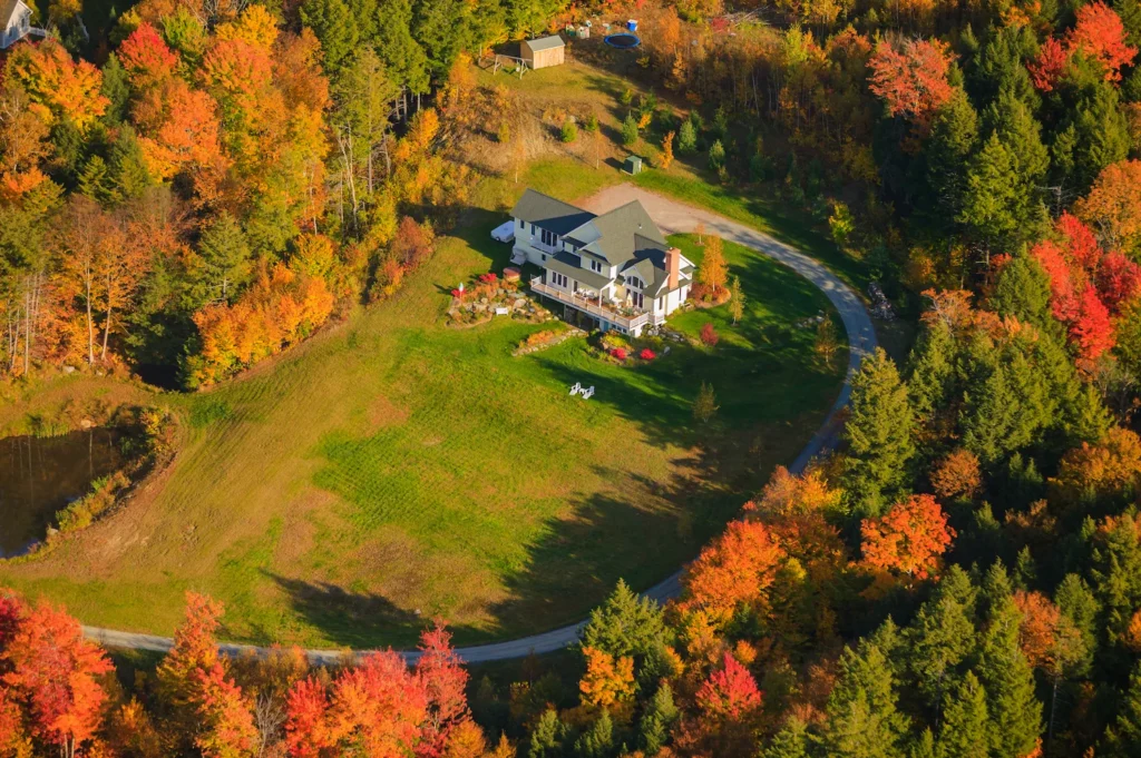 Aerial view of a house surrounded by a lawn and circular driveway, vibrant autumn trees in orange, red, and yellow bordering the property. A small pond and shed are visible—perfect for contemplating matters like disinheriting someone in Massachusetts.