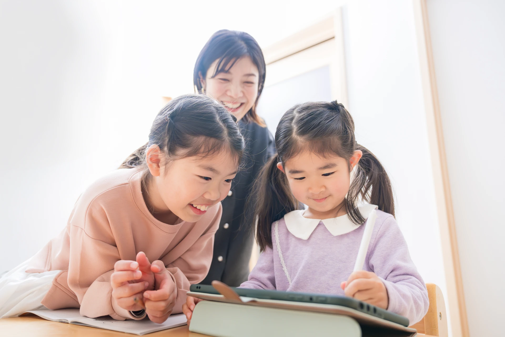 Two young girls use a tablet together, one writing with a stylus, while a smiling woman stands behind them, watching. They all appear happy and engaged in a bright, indoor setting.