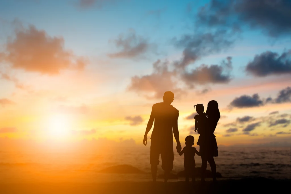 Silhouette of a family of four—two adults and two children—standing on the beach at sunset, with colorful clouds in the sky and the sun setting over the ocean in the background.