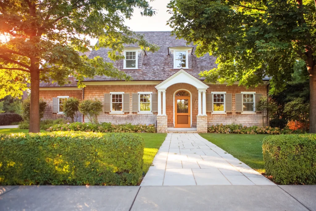 Charming brick house with a wooden front door, white columns, and dormer windows, surrounded by lush green trees, manicured bushes, and a stone walkway leading to the entrance on a sunny day.